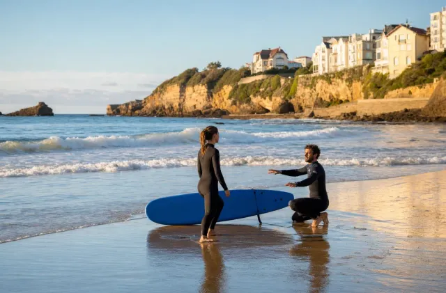 Moniteur de surf guidant une débutante avec une planche en mousse sur le sable mouillé à la Côte des Basques.